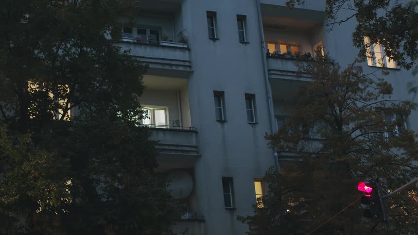 A static shot of a city apartment building at dusk, with lights glowing in the windows and a traffic light changing in the foreground