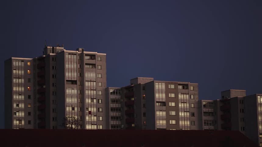 A still shot of urban apartment blocks with illuminated windows against a deep blue evening sky
