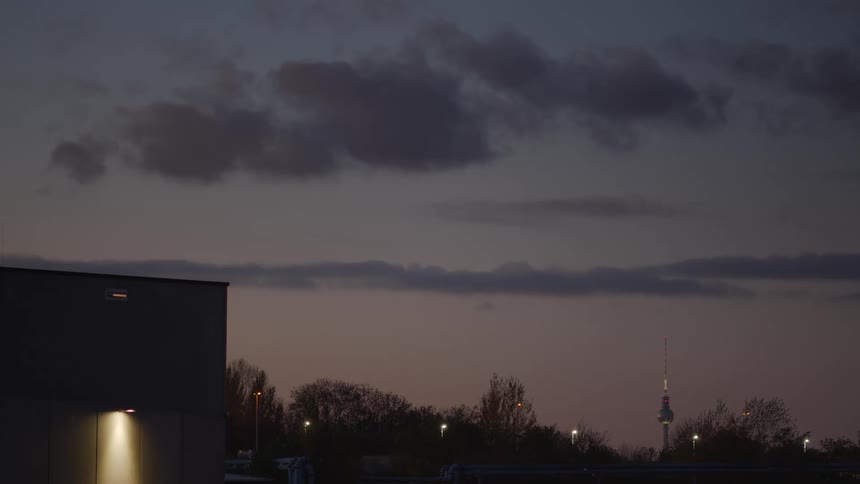 The iconic Berlin TV Tower is visible on the horizon at dusk, with a lit industrial building in the foreground under a cloudy sky