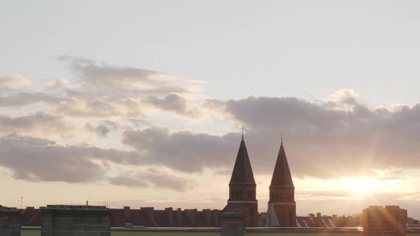 The sun sets behind the twin spires of a historic church, casting a warm glow over the city rooftops and cloudy sky
