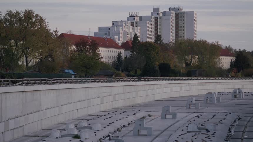 An empty, curving concrete floodway under construction contrasts with the residential buildings and trees of a city