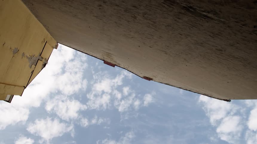 A worm's-eye view of clouds drifting across a blue sky, framed by weathered building walls in a narrow alley
