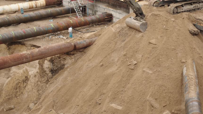 An excavator rests on a pile of sand at an industrial construction site with large metal pipes