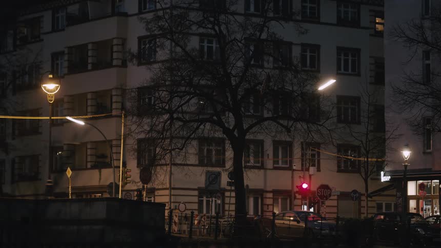 A static wide shot of a European city intersection at night with cars, bicycles, and pedestrians passing by