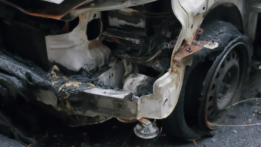 A detailed static shot shows the wreckage of a car fire, focusing on the charred front and melted rubber tire