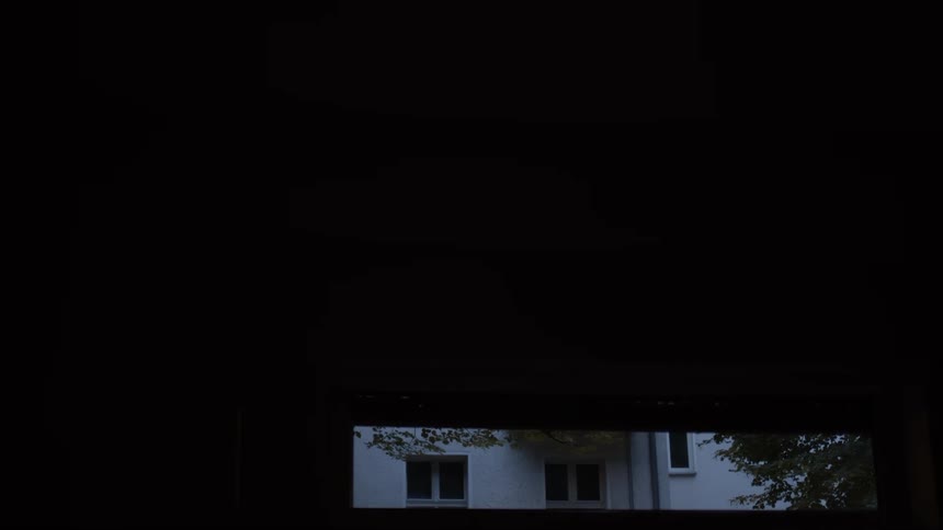 A black roller shutter slowly descends over a narrow window at dusk, gradually closing off the outside view and plunging the room into darkness