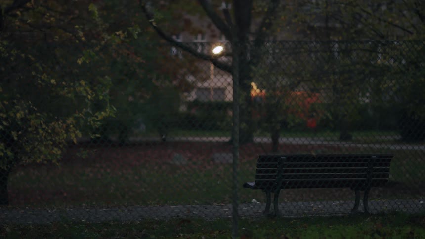 An empty park bench is seen through a chain-link fence on a quiet autumn evening as car lights pass in the distance