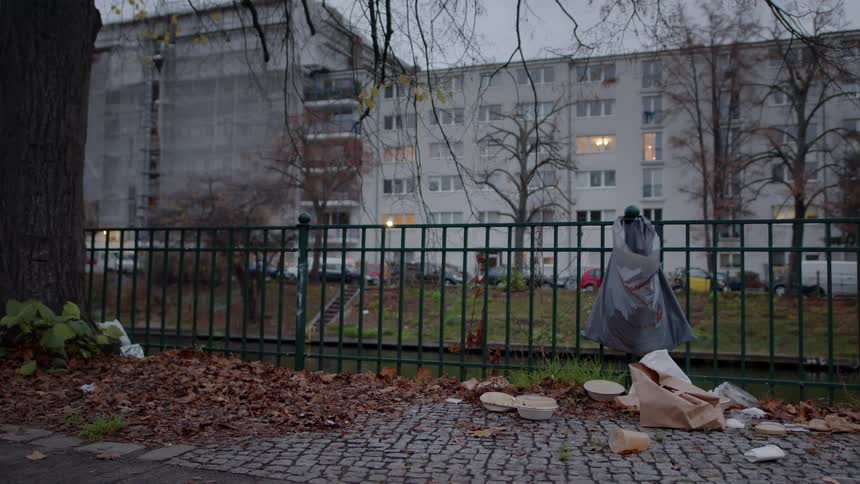 A bleak urban scene shows garbage littered on a cobblestone sidewalk next to a green fence during a dreary autumn day