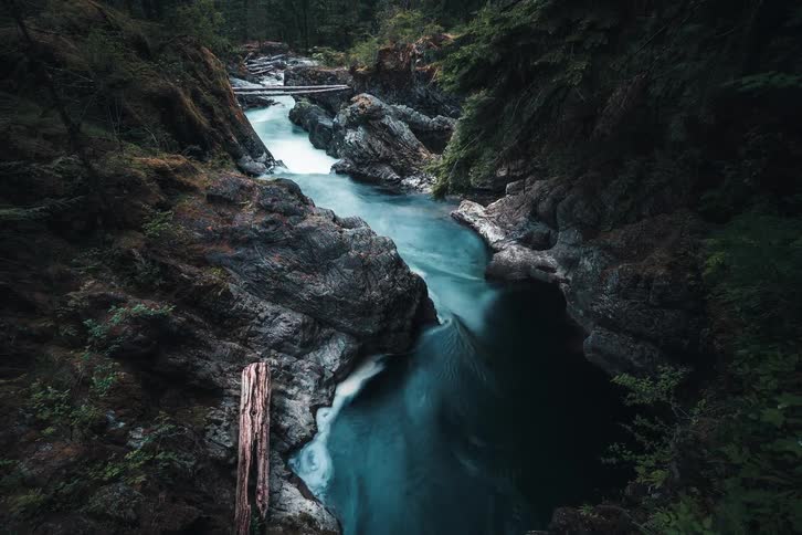 A dramatic long exposure shot of a turquoise river flowing through a dark and rocky forest canyon