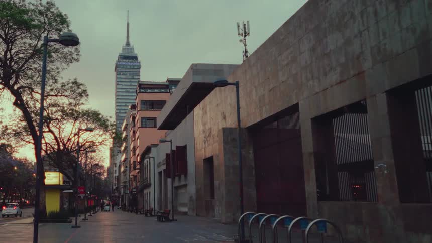 A cinematic dolly shot moves past a modern sculpture to reveal a quiet Mexico City street at dusk with the iconic Torre Latinoamericana in the background