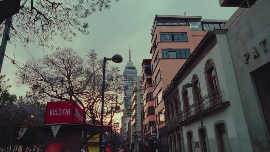 A slow dolly shot moves down a quiet street in Mexico City at dusk, revealing the iconic Torre Latinoamericana skyscraper in the distance