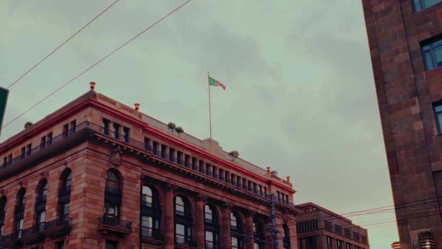 The Mexican flag proudly waves from the rooftop of a historic building in Mexico City under a moody, overcast sky