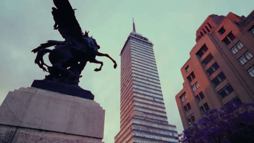 A low-angle, cinematic tilt-up shot reveals the Torre Latinoamericana skyscraper and a Pegasus statue against the dusk sky in Mexico City