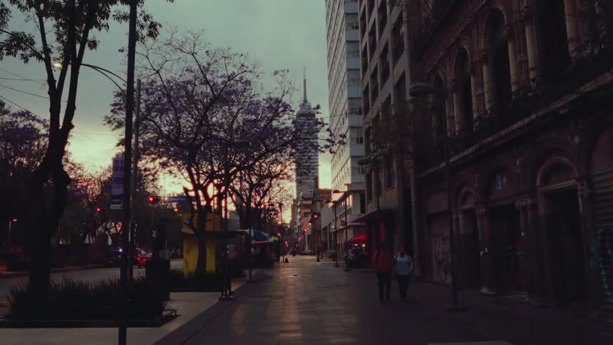 A slow push-in shot along a quiet city sidewalk in Mexico City at sunset, capturing the contrast between old buildings and a modern skyscraper in the distance