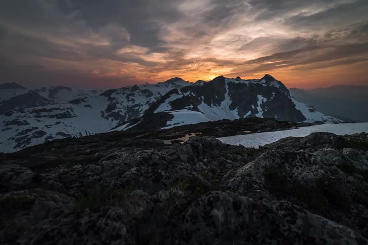 A dramatic timelapse captures the sun setting behind a vast, snow-covered mountain range as colorful clouds drift across the sky