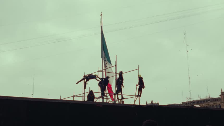 Silhouetted figures perform a slow-motion stunt fall from scaffolding in front of a Mexican flag against an overcast sky