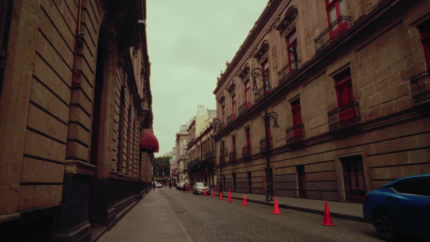 A slow dolly shot moves down a quiet, historic cobblestone street lined with ornate colonial-style buildings on an overcast day