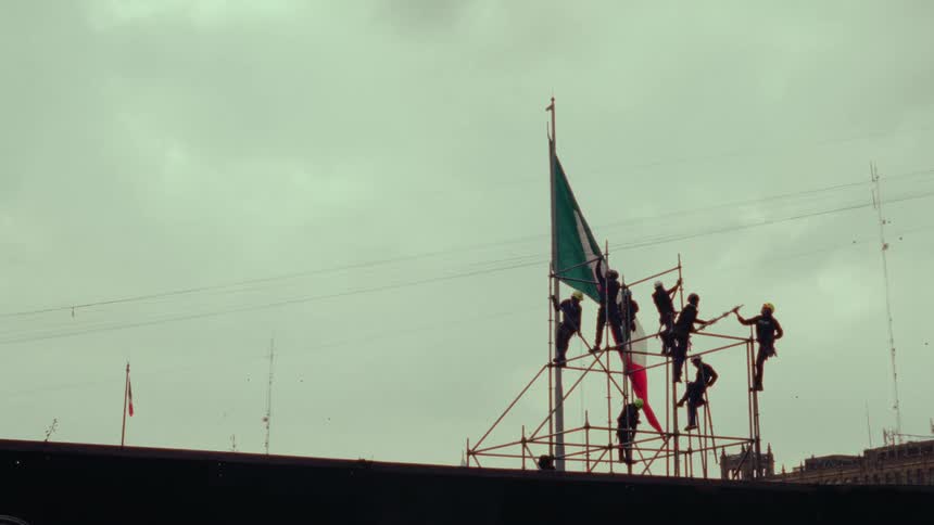 A group of men on scaffolding raise the Mexican flag against a cloudy gray sky