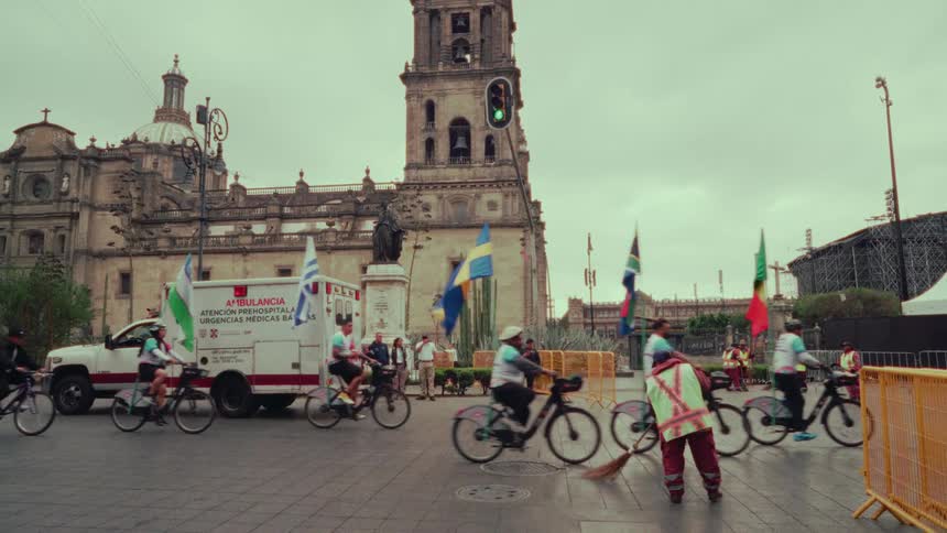 A group of bicyclists with various international flags ride through a historic city plaza as a worker sweeps the street