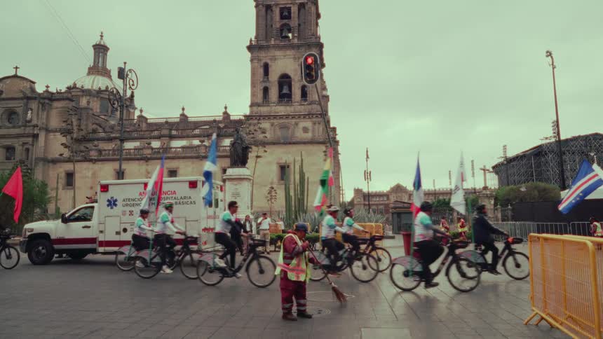 Cyclists carrying international flags participate in a parade in front of the Mexico City Metropolitan Cathedral as a street cleaner works