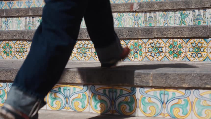 A person in jeans walks up historic stone stairs decorated with ornate, colorful ceramic tiles in slow motion