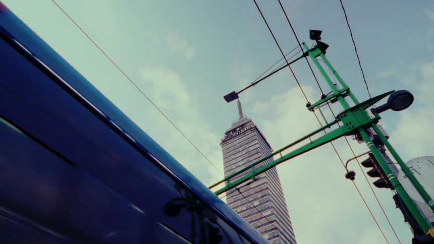 A low-angle shot follows a blue electric trolleybus as it passes by the Torre Latinoamericana in Mexico City