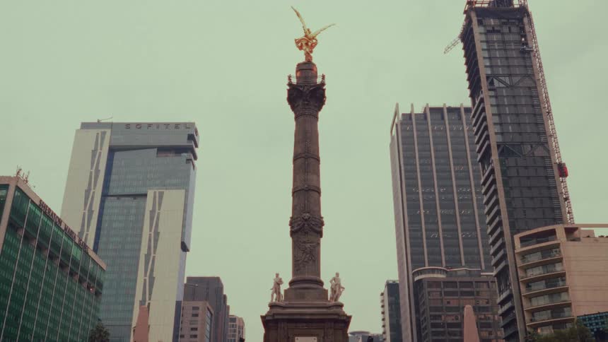 A static low-angle shot captures the historic Angel of Independence monument contrasting with the modern skyscrapers of Mexico City