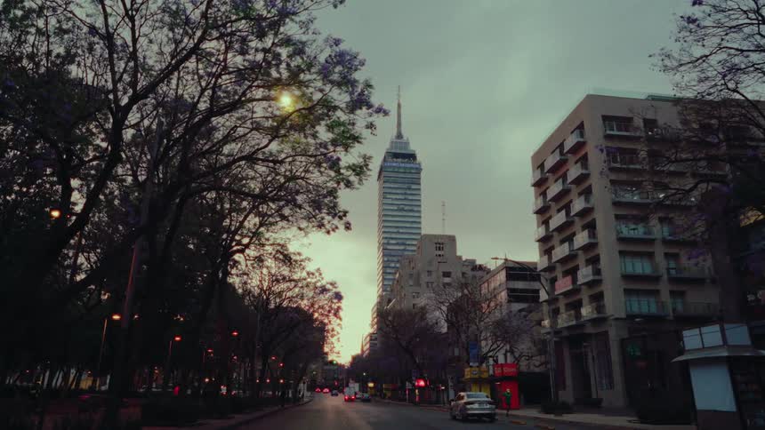 A slow forward dolly shot down a tree-lined street in Mexico City reveals the iconic Torre Latinoamericana skyscraper against a moody dusk sky
