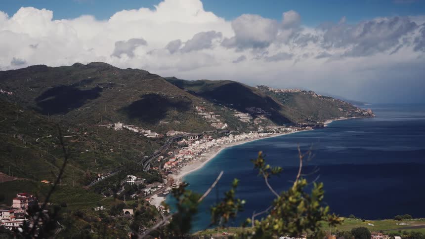 A stunning panoramic view of a seaside town along the Sicilian coast, nestled between lush green mountains and the deep blue sea under a partly cloudy sky