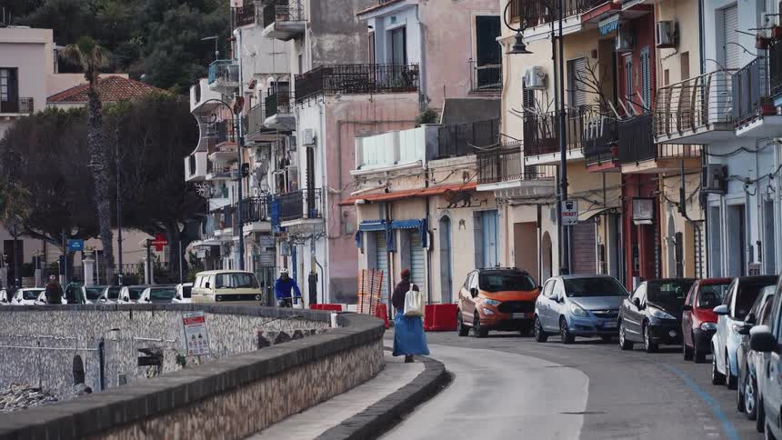 A woman in a long blue skirt walks down a curving seaside street in a quaint Italian town