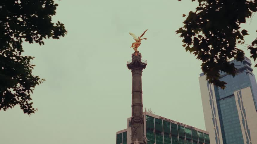A slow sideways pan reveals the iconic Angel of Independence monument in Mexico City from behind tree leaves