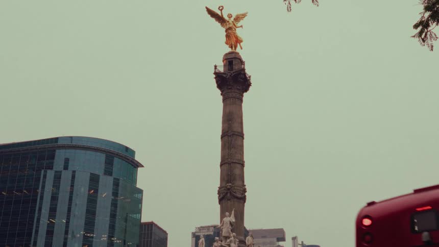 A red double-decker bus drives away, revealing the iconic Angel of Independence monument in Mexico City against a hazy sky