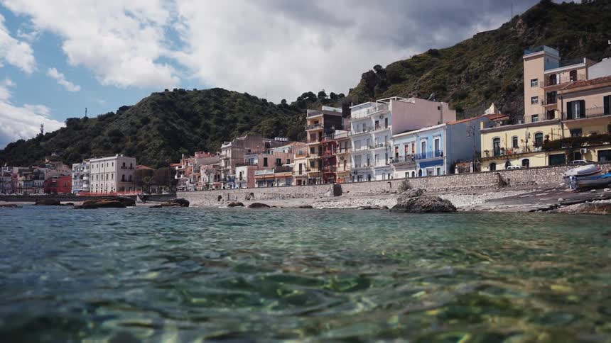 A serene water-level view of the colorful houses in a coastal town nestled at the base of a green hill in Italy