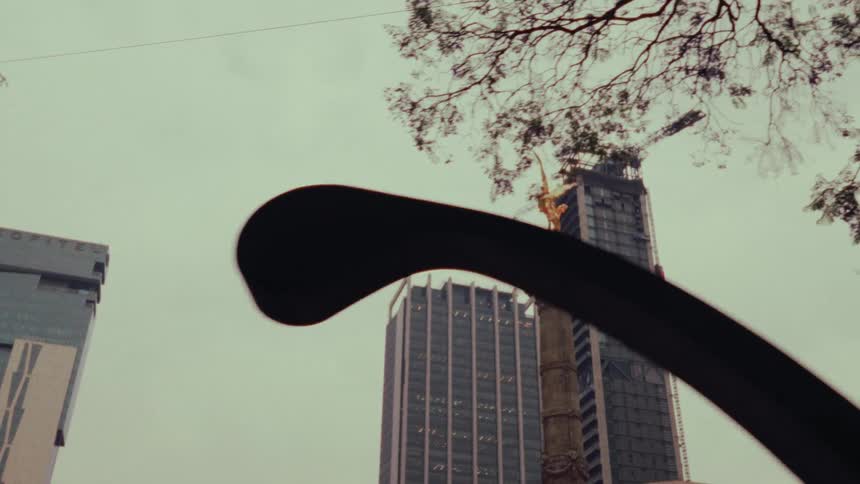 A slow dolly reveal of the Angel of Independence monument in Mexico City behind trees with modern skyscrapers in the background