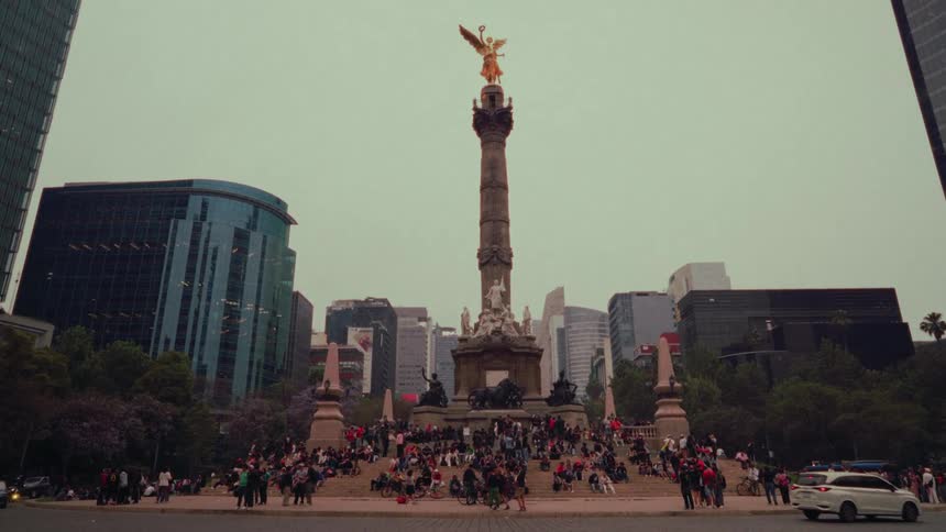 A slow tilt-up reveals the iconic Angel of Independence monument in Mexico City, where crowds gather against a backdrop of modern skyscrapers on an overcast day