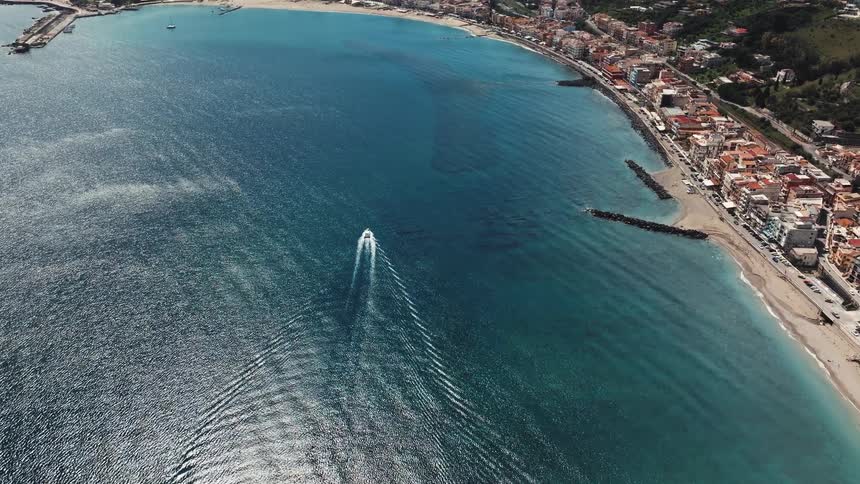 A stunning aerial view of a speedboat leaving a wake as it travels across the turquoise ocean towards a beautiful coastal town in Italy