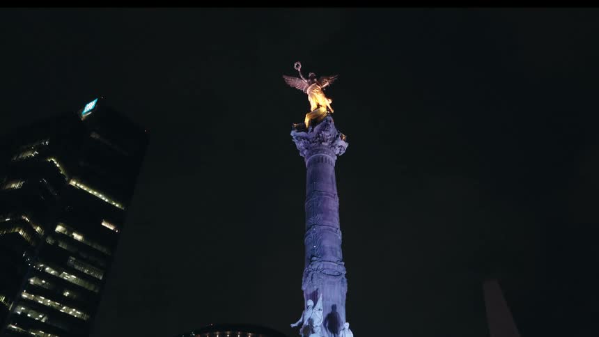The Angel of Independence monument in Mexico City is illuminated with purple and gold lights against the dark night sky