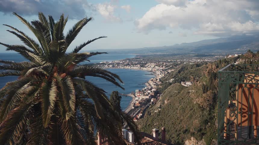 A stunning panoramic view from a balcony in Taormina, Sicily, shows the beautiful coastline and bay with a lush palm tree in the foreground