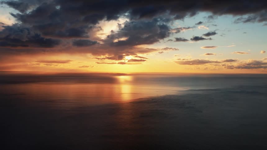 A serene and majestic aerial view shows a golden sunset reflecting on the calm ocean under a sky of dramatic, dark clouds