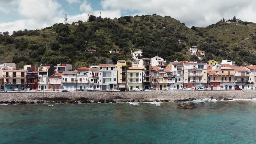 Scenic aerial footage pans across a charming Italian coastal town with colorful houses built along the turquoise shoreline at the base of a green hill