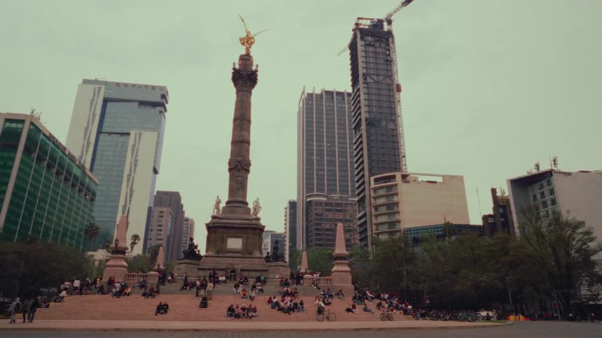 A wide panning shot of people gathered at the base of the Angel of Independence monument with the Mexico City skyline in the background