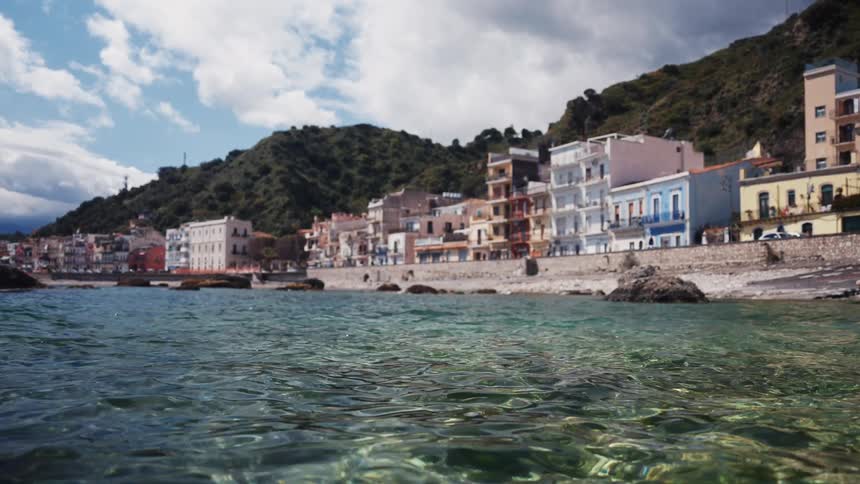 A scenic view from the clear, tranquil sea of a colorful coastal town nestled at the base of a verdant hill in Sicily, Italy
