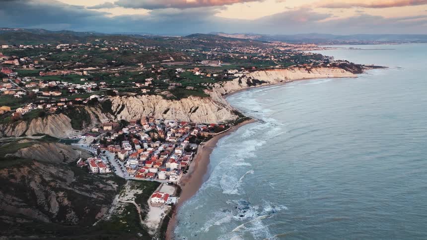 Static aerial view of a picturesque coastal village nestled beneath dramatic white cliffs along the Sicilian coastline at sunset