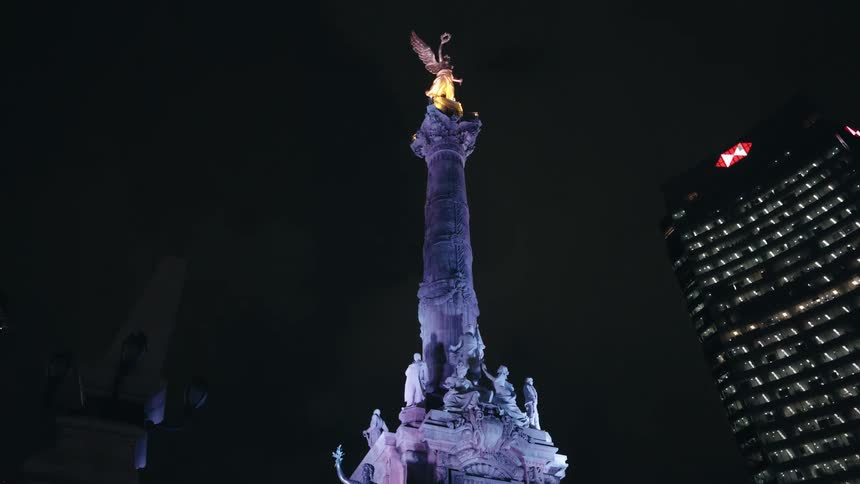A slow tilt up reveals the majestic Angel of Independence monument, illuminated by vibrant purple and gold lights against the dark Mexico City night sky