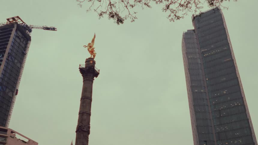 A low-angle pan reveals the iconic Angel of Independence monument standing tall amidst modern skyscrapers in Mexico City on an overcast day