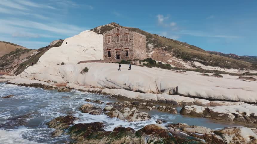 A stunning aerial view shows an abandoned stone building on the white rocky coast of Punta Bianca as waves crash on the shore
