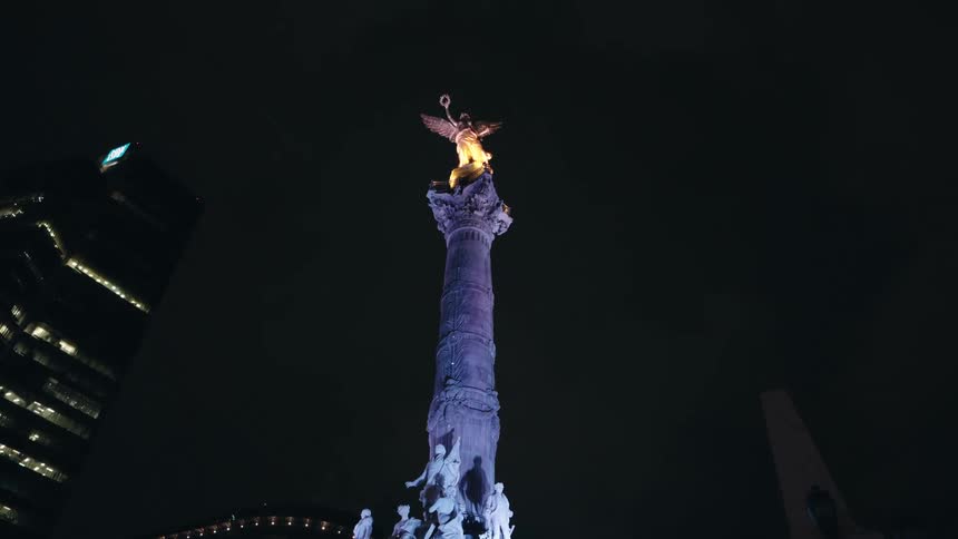 A low-angle view of the iconic Angel of Independence monument in Mexico City, illuminated with purple and gold lights against the dark night sky