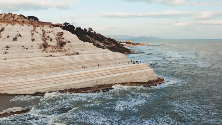 A majestic aerial view of the Scala dei Turchi in Sicily, where people walk along the brilliant white cliffs as waves crash below