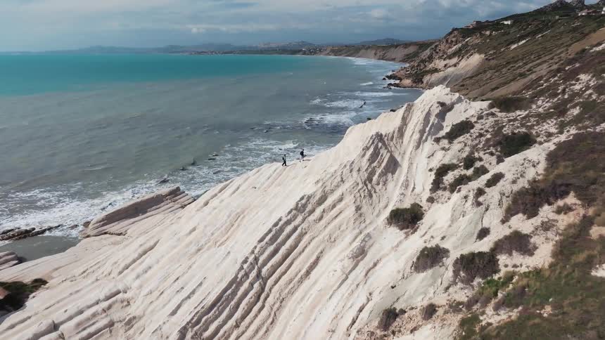 An aerial view shows two people walking along the stunning white cliffs of Scala dei Turchi in Sicily, with the turquoise sea below