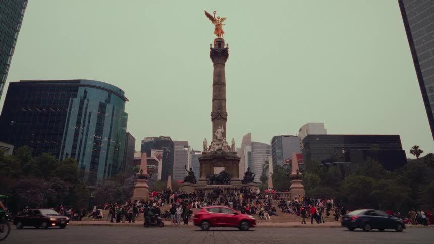 A wide, static shot captures the bustling traffic and crowds around the iconic Ángel de la Independencia monument in Mexico City